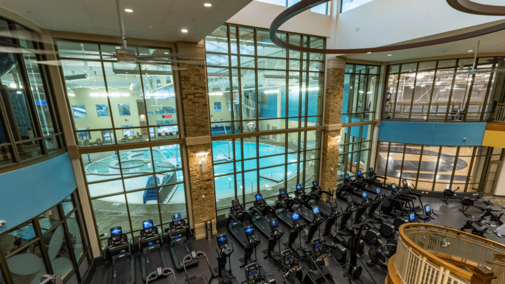 A spacious gym interior with a row of treadmills facing large windows. The windows overlook an indoor pool area with a turquoise circular pool. The space has high ceilings and modern architecture.